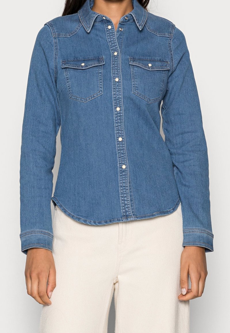 Femme portant une chemise en denim à manches longues boutonnée avec poches poitrine et pantalon blanc cassé, vue des épaules jusqu’aux hanches.