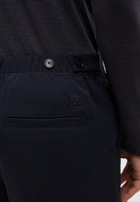 Gros plan sur la taille d'un pantalon marine avec des détails de bouton, un dos élastique et une poche horizontale, porté avec un haut à manches longues noir.