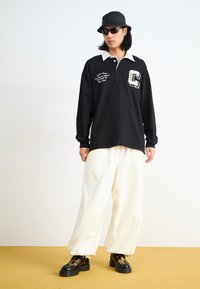 Black long-sleeve polo with white collar, featuring text detail and logo. Paired with loose white trousers and black boots. Yellow background.