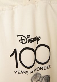 Crème stof met een bedrukte Disney-logo, "100," en "JAAR VAN VERRASSING" in het zwart. Inclusief een subtiele afbeelding van een pootafdruk.