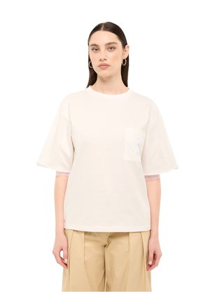 Woman with long dark hair wearing a loose white short-sleeve shirt and beige pants, standing against a plain white background.