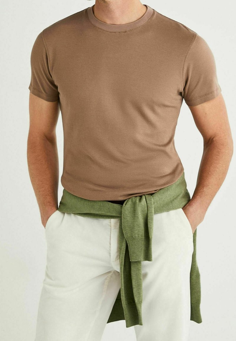 T-shirt marron à manches courtes côtelées avec un col rond, associé à un pull vert noué autour de la taille. Un pantalon blanc complète le look.