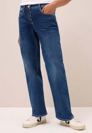 Jean droit bleu avec une fermeture à bouton, cinq poches et un léger délavage, associés à des baskets blanches avec un logo noir.