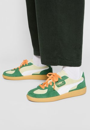 Stopy w zielono-beżowych sneakersach Puma Palermo z pomarańczowymi sznurówkami, białych skarpetkach i ciemnozielonych spodniach na jednolitym tle.
