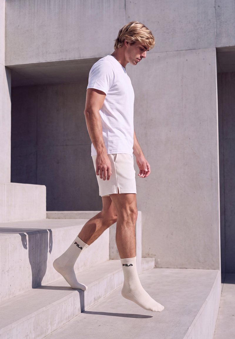 Jeune homme en t-shirt blanc, short et chaussettes FILA marchant pieds nus sur des marches en béton dans un cadre extérieur minimaliste.
