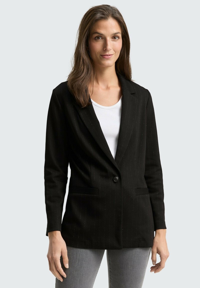 Zwarte blazer met krijtstreepje, enkelsluiting met knoop en ingesneden revers; gecombineerd met een wit shirt en grijze spijkerbroek. Soepele textuur.