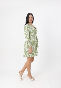 Vestido verde con estampado floral, de manga larga y con dobladillo en volantes. Llevado con sandalias de tacón blanco y con punta al aire. Fondo blanco liso.