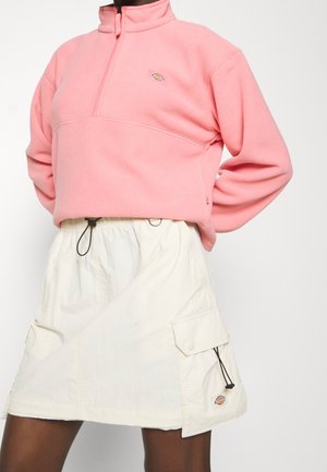 Lichtroze fleecetrekker met een halve ritssluiting en logopatch, gecombineerd met een witte rok met cargozakken en trekkoorddetails.