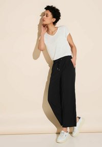 Kobieta w białej koszulce i czarnych szerokich spodniach opierająca się o beżową ścianę, jedną ręką dotykająca szyi, ubrana w białe sneakersy.