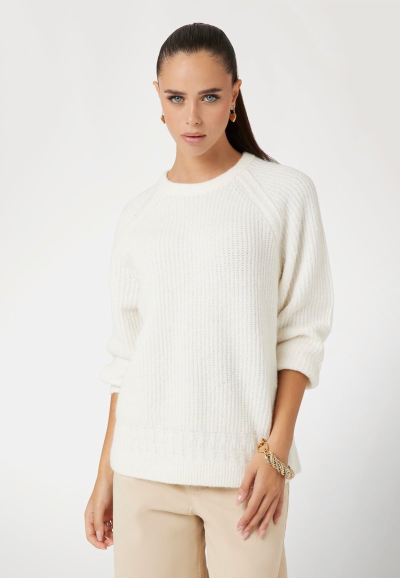 Pull en tricot couleur crème avec un motif en relief, col rond et manches trois-quarts. Porté avec un pantalon beige et un bracelet en or.