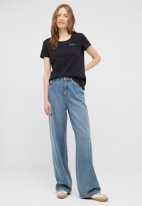 Czarna koszulka z krótkim rękawem o okrągłym dekolcie i białym logo. Połączona z luźnymi jasnoniebieskimi dżinsami oraz beżowymi butami.