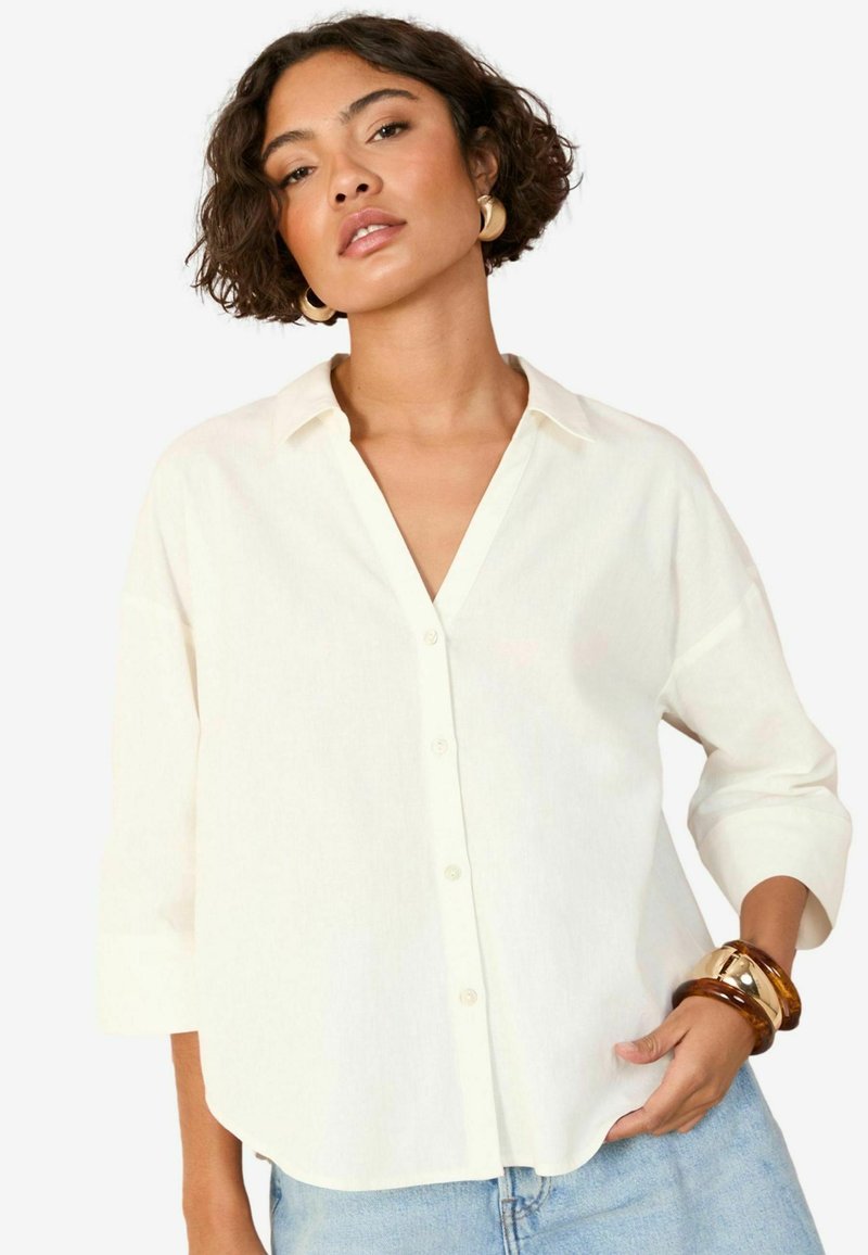 Camicia bianca a bottoni con scollatura a V, maniche a tre quarti e orlo curvo; tessuto leggero con una consistenza morbida. Bracciali in legno e metallo.