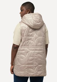 Hellbeige gepolsterte Weste mit Kapuze, die ein gestepptes Muster und seitliche Taschen aufweist, getragen über einem cremefarbenen Strickoberteil.