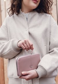 Pink leather wallet with a zip closure, featuring a silver logo and a carrying strap, held against a soft beige sweatshirt.