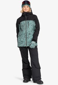 Sneeuwjack in teal met zwarte accenten, voorzien van een bloemenpatroon, een ritssluiting aan de voorkant, twee zakken, gedragen met zwarte snowboardbroeken en handschoenen.