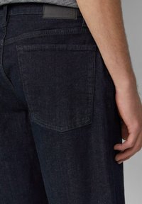 Jean en denim bleu foncé avec une coupe slim, doté d'une poche arrière et d'une étiquette en cuir. La texture est lisse avec un léger éclat.