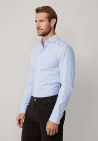Camisa azul claro de manga larga con cuello casual y botonadura en el frente, hecha de una tela con una textura suave. Presenta un pequeño logo y puños.
