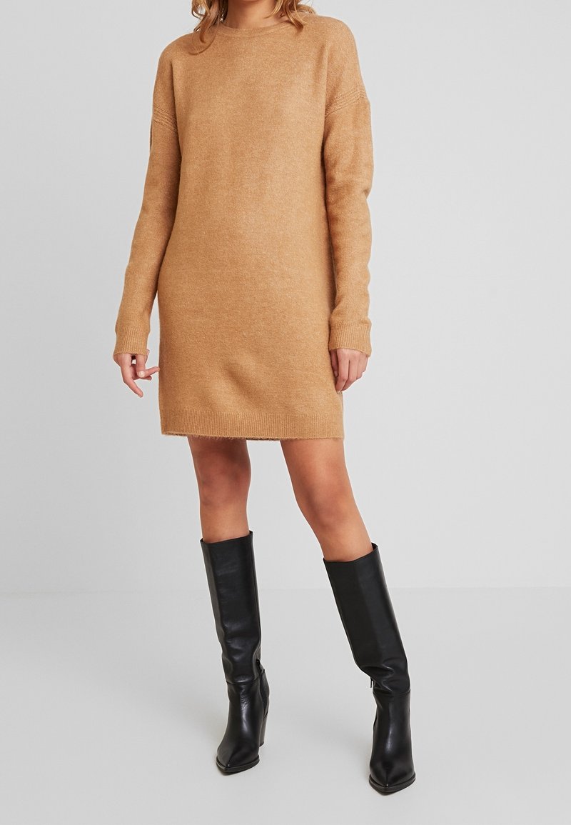 Robe pull en maille de couleur marron clair, à manches longues et coupe décontractée. Portée avec des bottes en cuir noir montant jusqu'aux genoux. Texture lisse, détails minimalistes.