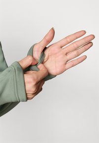 Grønn langermet topp med tommelhull-funksjon. Stoffet er mykt og elastisk, og viser en hånd som justerer ærmet rundt tommelen.