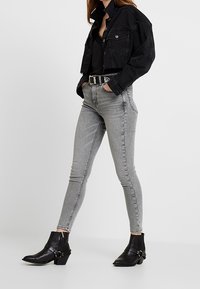 Chaqueta de mezclilla negra corta sobre una blusa negra, combinada con pitillos grises y botas negras al tobillo con acentos de tachuelas.