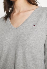 Grå ribbet v-hals sweater med et lille broderet logo i rød, hvid og blå, som fremhæver en afslappet pasform og blød tekstur.