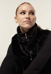 Musta tekoturkisliivi korkean kauluksen, etuvetoketjun ja sileän pinnan kanssa. Malli pukeutunut mustaan pitkähihaiseen puseroon; neutraali tausta.