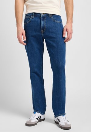 Mann trägt blaue Jeans mit geradem Bein und weiße Sneakers mit schwarzen Streifen, steht vor einem einfarbigen hellen Hintergrund.