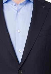 Granatowa marynarka o teksturowanej fakturze, z wyłogami i jednym guzikiem, zestawiona z jasnoniebieską koszulą z kołnierzykiem, posiadającą białe guziki.