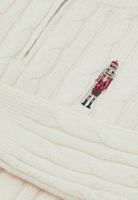 Fehér kötött pulóver kábelmintával és félcipzárral ellátott gallérral. A mellrészen egy kis hímzett diótörő dísz található.