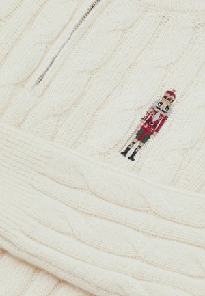 Fehér kötött pulóver kábelmintával és félcipzárral ellátott gallérral. A mellrészen egy kis hímzett diótörő dísz található.