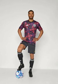 Meeste lühikeste varrukatega spordisärk punases ja tumepunasest geomeetrilises mustris, kombineeritud hallide lühikeste pükste, mustade sokkide ja jalgpalliketsidega, koos palliga.