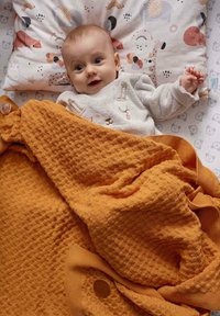 Pomarańczowy teksturowany koc okrywa niemowlę na wzorzystej poduszce. Koc ma dzianinowy wzór, kontrastujący z miękkim materiałem.