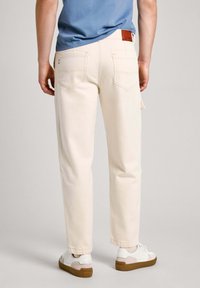 Lys beige bukser med lige snit, baglommer og en brun læderlabel. Bæres med hvide sneakers og en blå skjorte. Glat tekstur.