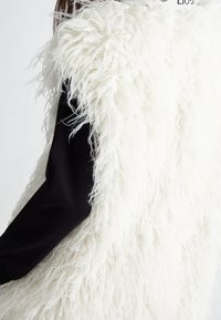 Fluffy weißer Kunstfell-Weste mit strukturiertem Finish, kombiniert mit einem figurbetonten schwarzen Langarmoberteil. Merkmale einen runden Halsausschnitt und eine offene Vorderseite.