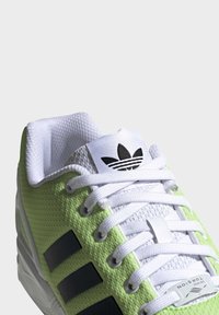 Green mesh sneaker with white laces, black stripes, and a white tongue featuring a black logo. Detailed texture on upper surface.
