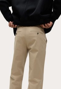Beige chino's met achterzakken en knoopsluitingen. Materiaal lijkt glad met een getailleerde pasvorm, gedragen met een zwart sweatshirt.