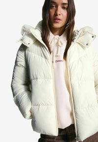 Jonge vrouw die een crèmekleurige pufferjack met capuchon en een witte hoodie met trekkoorden draagt, staande tegen een effen witte achtergrond.