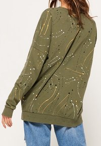Sudadera verde oliva con patrones abstractos en amarillo, blanco y negro; cuello redondo; mangas largas; corte relajado; puños y dobladillo acanalados.