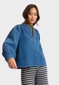 Denim-Oberteil mit tiefem V-Ausschnitt und Puffärmeln, mit einer lockeren Passform und genähten Akzenten. Kombiniert mit gestreiften, mehrfarbigen Hosen.