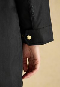 Schwarzer wasserdichter Jackenärmel mit einem goldenen Druckknopf, auf dessen Seite "JOULES" in subtil geprägter Schrift gedruckt ist.