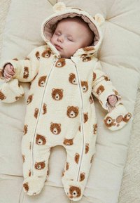 Bambino che dorme vestito con un morbido tutù crema con facce di orsetto marroni e cappuccio con orecchie da orso, sdraiato su un cuscino beige.