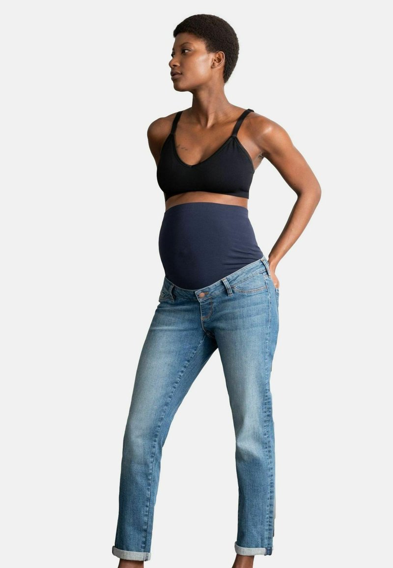 Femme enceinte portant un soutien-gorge de maternité noir et un jean de maternité bleu, debout avec les mains reposant sur les hanches, regardant sur le côté.