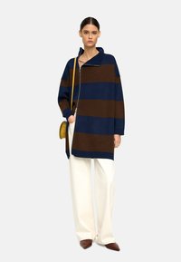 Przesadna wełniana kurtka w połączeniu z włóknami sztucznymi w niebieskie i brązowe paski, z zamkiem błyskawicznym i wysokim kołnierzem. W połączeniu z szerokimi białymi spodniami.