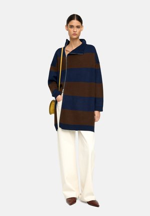 Przesadna wełniana kurtka w połączeniu z włóknami sztucznymi w niebieskie i brązowe paski, z zamkiem błyskawicznym i wysokim kołnierzem. W połączeniu z szerokimi białymi spodniami.