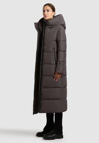 Long puffer coat in dark gray with a high collar, zippered front, side pockets, and a quilted design. Paired with black pants and boots.