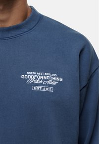 Niebieska bluza z prążkowanym kołnierzem i haftowanym napisem: "Północno-Zachodnia Anglia, Good For Nothing, British Atelier, założona w 2011."