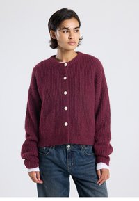 Burgundy knitted cardigan with a crew neckline, featuring large white buttons and ribbed texture; layered over a white long-sleeve top.