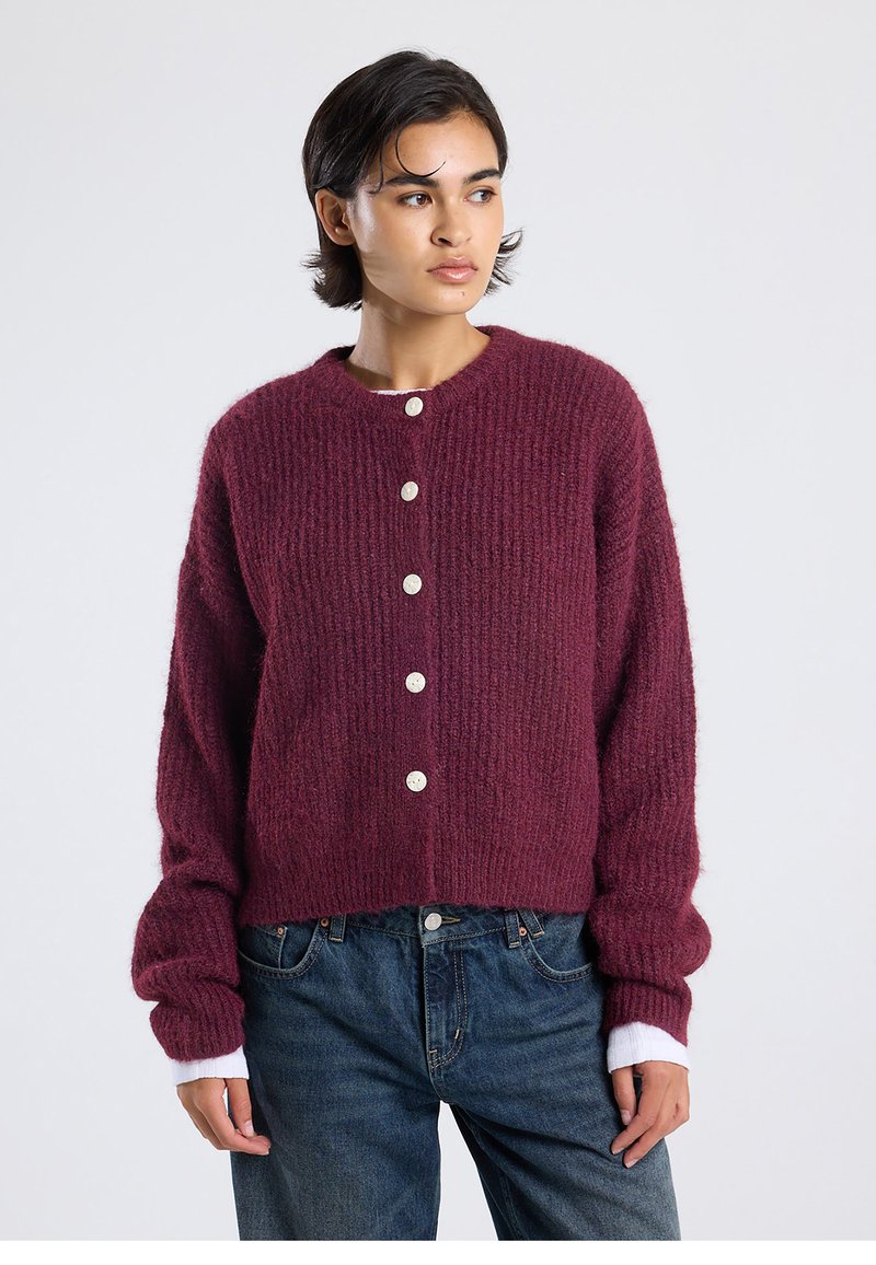 Burgundy knitted cardigan with a crew neckline, featuring large white buttons and ribbed texture; layered over a white long-sleeve top.