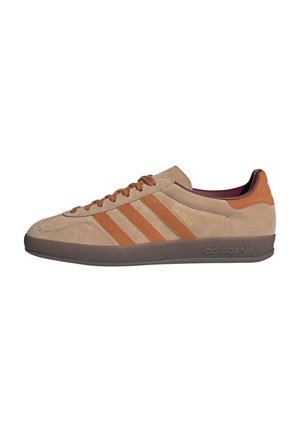 Baskets Adidas Gazelle en daim beige avec des bandes marron, des lacets marron, une semelle foncée et un accent orange au talon, présentées de profil sur fond blanc.