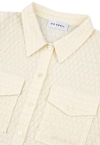 Chemise couleur crème en tissu texturé. Elle bénéficie d'un col, de deux poches à rabat et de boutons en perles. L'étiquette indique "OH APRIL".